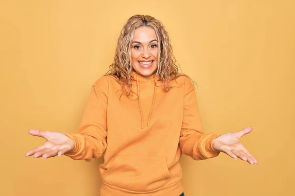 stock image Young beautiful blonde sporty woman wearing casual sweatshirt over yellow background smiling cheerful offering hands giving assistance and acceptance.