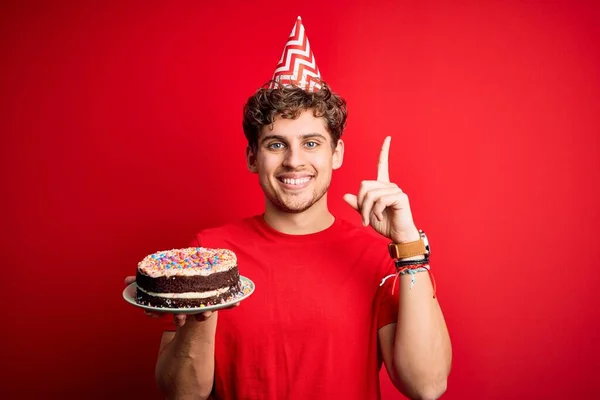 Young blond man with curly hair wearing birthday hat holding cake over red background surprised with an idea or question pointing finger with happy face, number one