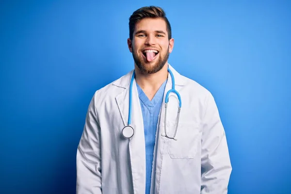 ひげと青い目をした若いブロンドの医者男が白いコートと聴診器を身に着けていて面白い表情で幸せに舌を突き出しています 感情概念 — ストック写真
