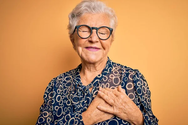 Senior beautiful grey-haired woman wearing casual shirt and glasses over yellow background smiling with hands on chest with closed eyes and grateful gesture on face. Health concept.