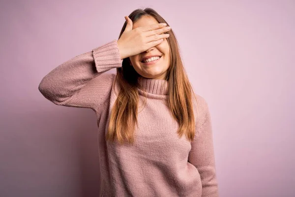 Beautiful young woman wearing turtleneck sweater over pink isolated background smiling and laughing with hand on face covering eyes for surprise. Blind concept.