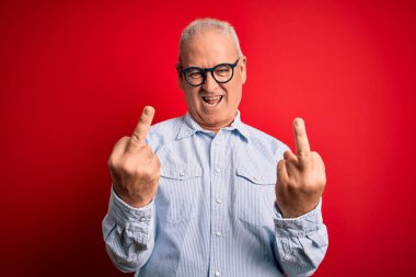Middle age handsome hoary man wearing casual striped shirt and glasses over red background Showing middle finger doing fuck you bad expression, provocation and rude attitude. Screaming excited