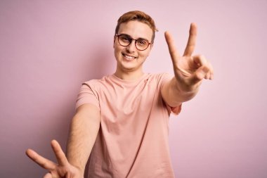 Genç, kızıl saçlı, günlük tişört giyen, pembe arka planda dikilen ve iki elini de zafer işareti yaparken gösteren bir gülümseme ile gülümseyen genç adam. İki numara..