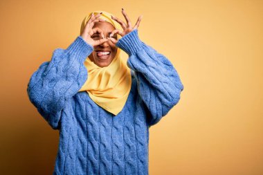 İzole edilmiş sarı arka planda Müslüman tesettürlü genç Afrikalı Afro kadın dürbünün dili dışarı çıkarması, gözlerin parmaklarından içeri bakması gibi güzel hareketler yapıyor. Çılgın ifade.