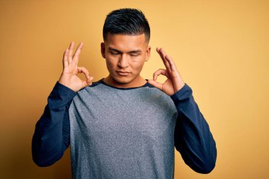 Sarı arka planda dikilen gündelik tişört giyen genç, yakışıklı Latin adam rahatla ve gözün kapalı gülümse parmaklarınla meditasyon hareketi yap. Yoga konsepti.