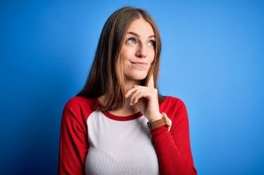 Genç, güzel, kızıl saçlı bir kadın izole edilmiş mavi arka plan üzerine gündelik tişört giyiyor. Elleri çenesinde soru sormayı düşünüyor. Düşünceli bir ifade. Düşünceli bir yüzle gülümsüyor. Şüphe kavramı.