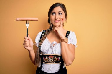 Genç esmer Alman kadın geleneksel Ekim Festivali elbisesi giyiyor. Elinde sosis var. Ciddi bir yüz ifadesi var.