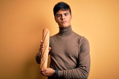 Genç yakışıklı adam, izole edilmiş sarı arka planda taze ekmek tutuyor. Akıllı bir yüz ifadesiyle ciddi ciddi düşünüyor.