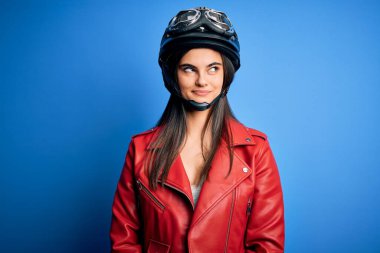 Motosiklet kaskı ve ceketi giyen, gülümseyen ve gözlerini kaçıran güzel, esmer, motosikletli bir kadın..