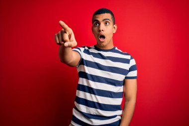 Yakışıklı Afro-Amerikan erkeği kırmızı arka planda dikilen sıradan çizgili tişört giyiyor. Parmakları önde, ağzı açık ve şaşırmış bir şekilde işaret ediyor.
