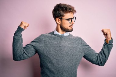 Genç, sakallı, gözlüklü ve süveterli, pembe arka planda duran yakışıklı bir adam gururla gülümseyen kol kaslarını gösteriyor. Fitness konsepti.