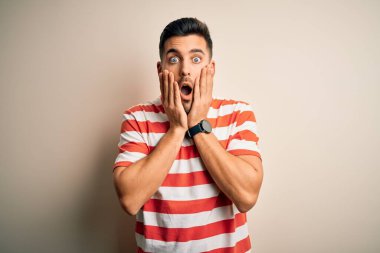 Sıradan çizgili tişört giyen genç yakışıklı adam izole edilmiş beyaz arka planda duruyor korkmuş ve şok olmuş, şaşırmış ve şaşırmış bir yüz ifadesi var.