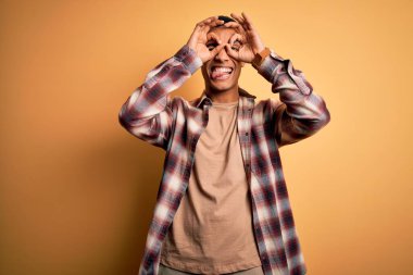 Genç, yakışıklı, Afro-Amerikalı bir adam sarı arka plan üzerinde dikilmiş dürbün gibi jest yapıyor, gözleri parmaklarından bakıyor. Çılgın ifade.