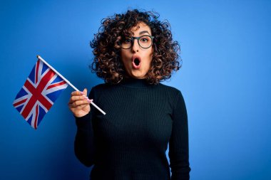 Genç, kıvırcık Arap kadın mavi arka planda İngiliz bayrağı tutan gözlüklü. Şaşırmış bir yüzle korkmuş ve korku ifadesinden heyecanlanmış.