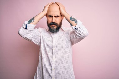 Sakallı yakışıklı kel bir adam. Soyutlanmış pembe arka planda zarif bir gömlek giyiyor. Ağrı ve migren yüzünden baş ağrısı çekiyor. Ellerini başının üstüne koy..