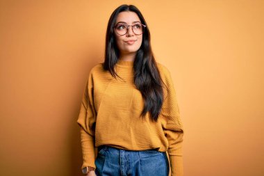 Genç esmer kadın gözlük takıyor ve sarı izole edilmiş arka planda rahat bir süveter giyiyor. Gülümsüyor ve uzaklara bakıyor..