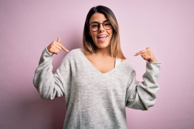 Genç, güzel esmer kadın pembe arka planda rahat süveter ve gözlük takıyor. Kendine güler yüzle bakıyor, kendini gururlu ve mutlu bir şekilde gösteriyor..