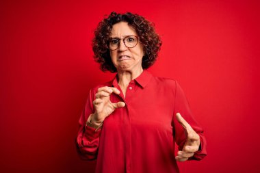 Orta yaşlı, kıvırcık saçlı, sıradan gömlekli, kırmızı arka planda gözlüklü bir kadın tiksindirici ve tiksindirici bir yüz ifadesi takınıyor..