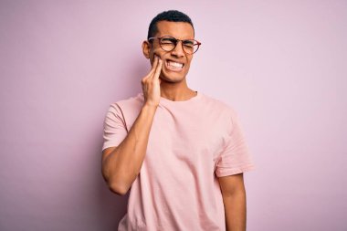 Yakışıklı Afro-Amerikan erkeği, günlük tişört giyip pembe arka plan üzerine gözlük takıyor diş ağrısı ya da diş ağrısı yüzünden ağıza acı dolu bir ifadeyle dokunuyor. Dişçi