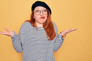 Sarı arka planda çizgili tişört ve Fransız beresi takan güzel kızıl saçlı kadın habersiz ve elleri ve kolları havada şaşkın bir ifade sergiliyor. Şüphe kavramı.
