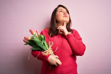 Güzel, iri yarı bir kadın pembe arka planda romantik lale demetleri tutuyor. Şüpheye odaklanmış, parmakları çenesinde ve yukarı bakıyor.