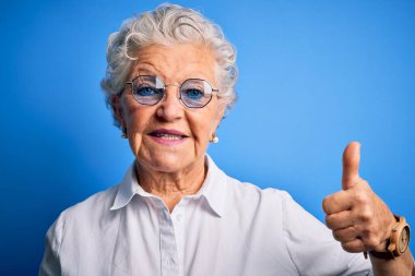 Üst düzey, zarif gömlek ve gözlük takan güzel bir kadın izole edilmiş mavi arka planda mutlu bir el hareketi yapıyor. Kameraya bakıp başarıyı gösteren ifade onaylanıyor.