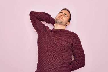 Mavi gözlü, sıradan bir süveter giyen, pembe arka planda duran, boynu ağrıyan, eliyle boynuna dokunan, kaslı bir adam.