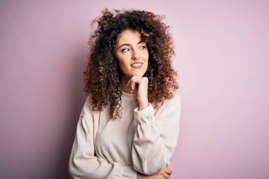 Kıvırcık saçlı ve piercingli güzel bir kadın pembe arka planda sıradan bir süveter giyiyor, elleri çenesinde soru sormayı düşünüyor, düşünceli bir yüz ifadesi var. Düşünceli bir yüzle gülümsüyor. Şüphe kavramı.