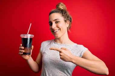 Güzel sarışın kadın kırmızı zemin üzerinde ferahlamak için kola köpüklü içecek içiyor. Elinle ve parmağınla işaret ederek çok mutlu.