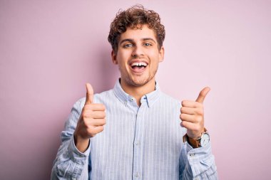 Sarışın, kıvırcık saçlı, beyaz arka plan işareti üzerine çizgili gömlek giyen, el hareketleriyle olumlu hareket eden, gülümseyen ve mutlu genç bir adam. Neşeli ifade ve kazanan jest.