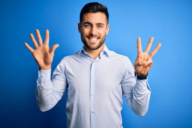 Zarif gömlekli genç adam mavi arka plan üzerinde duruyor ve 8 numaralı parmaklarıyla işaret ediyor kendinden emin ve mutlu bir şekilde gülümsüyor..