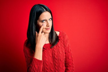 Mavi gözlü genç esmer kadın izole edilmiş kırmızı arka plan üzerine günlük süveter giyiyor. Gözünü dikip jestini izliyor. Şüpheli bir ifade.