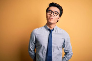 Genç yakışıklı Çinli iş adamı gözlük takıyor ve sarı arka planda kravat takıyor. Gülümsüyor ve başka tarafa bakıyor..