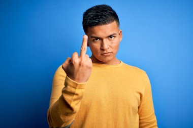 Young handsome latin man wearing yellow casual sweater over isolated blue background Showing middle finger, impolite and rude fuck off expression