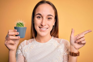 Güzel bir kadın, sarı arka planda küçük bir kaktüs saksısı tutuyor. Elleri ve parmakları kenarda mutlu bir şekilde işaret ediyor.