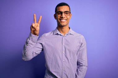 Yakışıklı Afro-Amerikalı, çizgili gömlek ve gözlük takmış mor arka planda gülümseyen ve parmaklarını zafer işareti yaparken gösteren kameraya bakan adam. İki numara..