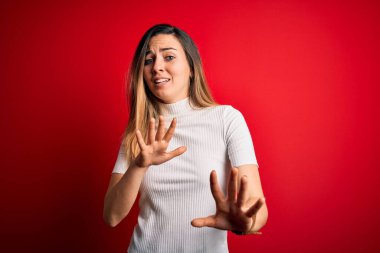 Kırmızı arka planda sıradan beyaz tişört giyen, mavi gözlü güzel sarışın kadın korku ifadesinden korkmuş ve dehşete düşmüş. El hareketlerini bırak, şok içinde bağır. Panik konsepti.