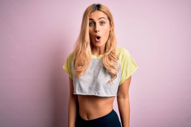 Genç, güzel, sarışın, spor yapan, spor giyinen, pembe arka planda giyinen, korku ve heyecan dolu bir yüz ifadesinden korkan ve şok olan genç bir kadın..