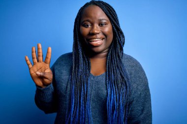 Mavi arka plan üzerine günlük süveter giyen Afrikalı Amerikalı artı cüsseli kadın parmaklarını dört numarayı gösterirken kendinden emin ve mutlu bir şekilde gülümsüyor..