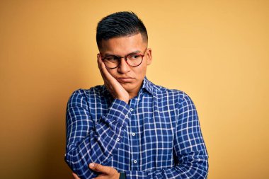 Sarı arka plan üzerinde gündelik gömlek ve gözlük takan genç, yakışıklı Latin adam kollarını kavuşturarak depresyondan sıkılmış ve yorgun görünüyor..