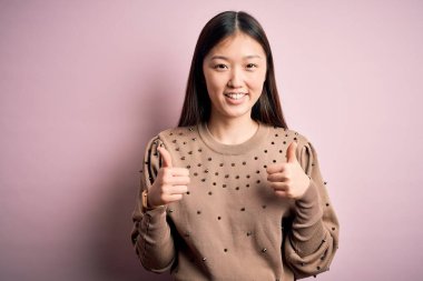 Güzel Asyalı bir kadın pembe arka plan başarı tabelasının üzerine şık bir süveter giyip el hareketleriyle olumlu jest yapıyor. Gülümsüyor ve mutlu. Neşeli ifade ve kazanan jest.