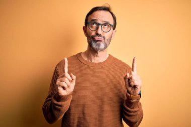 Kahverengi süveter ve gözlük takan orta yaşlı beyaz bir adam izole edilmiş sarı arka plan üzerinde üzgün ve üzgün görünüyor, parmaklarıyla yönü gösteriyor, mutsuz ve depresif..