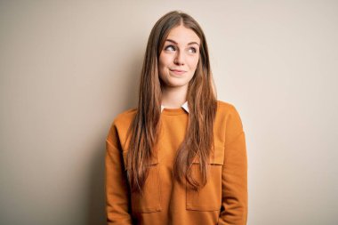 Genç, güzel, kızıl saçlı bir kadın izole edilmiş beyaz bir arka plan üzerine günlük süveter giyiyor. Gülümsüyor ve başka tarafa bakıyor..