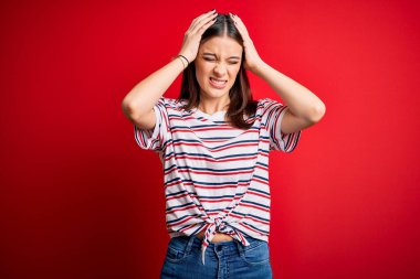 Genç ve güzel esmer bir kadın, günlük çizgili tişört giyiyor. Kırmızı arka planda dikiliyor. Ağrı ve migrenden dolayı çaresiz ve stresli. Ellerini başının üstüne koy..