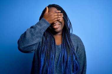 Mavi arka planda günlük süveter giyen Afrikalı Amerikalı artı cüsseli bir kadın. Gülümsüyor ve elleri yüzünde şaşkınlıktan gözleri kapayarak gülüyor. Kör kavram.