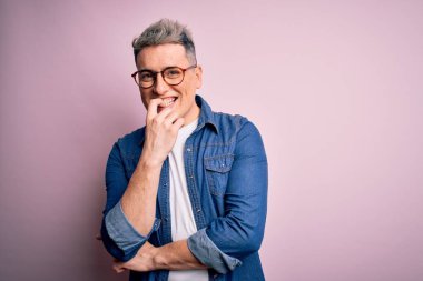 Pembe izole edilmiş arka planda gözlük ve kot ceket giyen genç ve modern bir adam kameraya kollarını kavuşturup gülümseyerek ve çenesini kaldırarak bakıyor. Olumlu düşünüyorum..