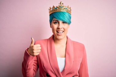 Mavi saçlı, pembe izole edilmiş arka planda kraliçe tacı takan genç bir iş kadını mutlu bir el hareketi yapıyor. Kameraya bakıp başarıyı gösteren ifade onaylanıyor.