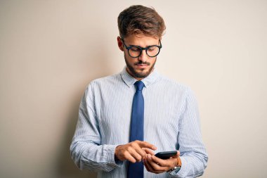 Genç iş adamı, akıllı telefonu beyaz arka plan üzerinde kullanıyor. Akıllı bir yüz ifadesiyle ciddi ciddi düşünüyor.