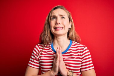 İzole edilmiş kırmızı arka plan üzerinde gündelik çizgili tişört giyen genç ve güzel sarışın kadın yüzünde umut ifadesiyle birlikte yalvarıyor ve dua ediyor. Çok duygusal ve endişeli. Yalvarıyorum..