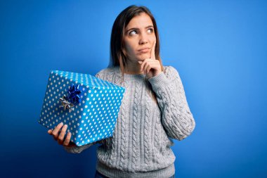 Sarışın genç bir kadın, izole edilmiş arka planda doğum günü hediyesi olarak hediye kutusunu tutuyor. Ciddi bir yüz. Soru sormayı düşünüyor.